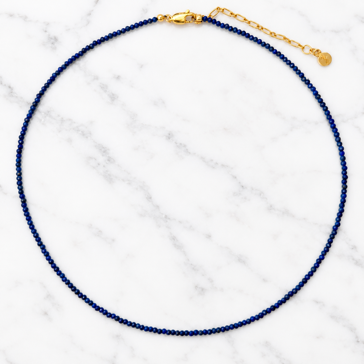 A beaded necklace made of small dark blue lapis beads with a gold clasp and extender chain, displayed on a white marble background.