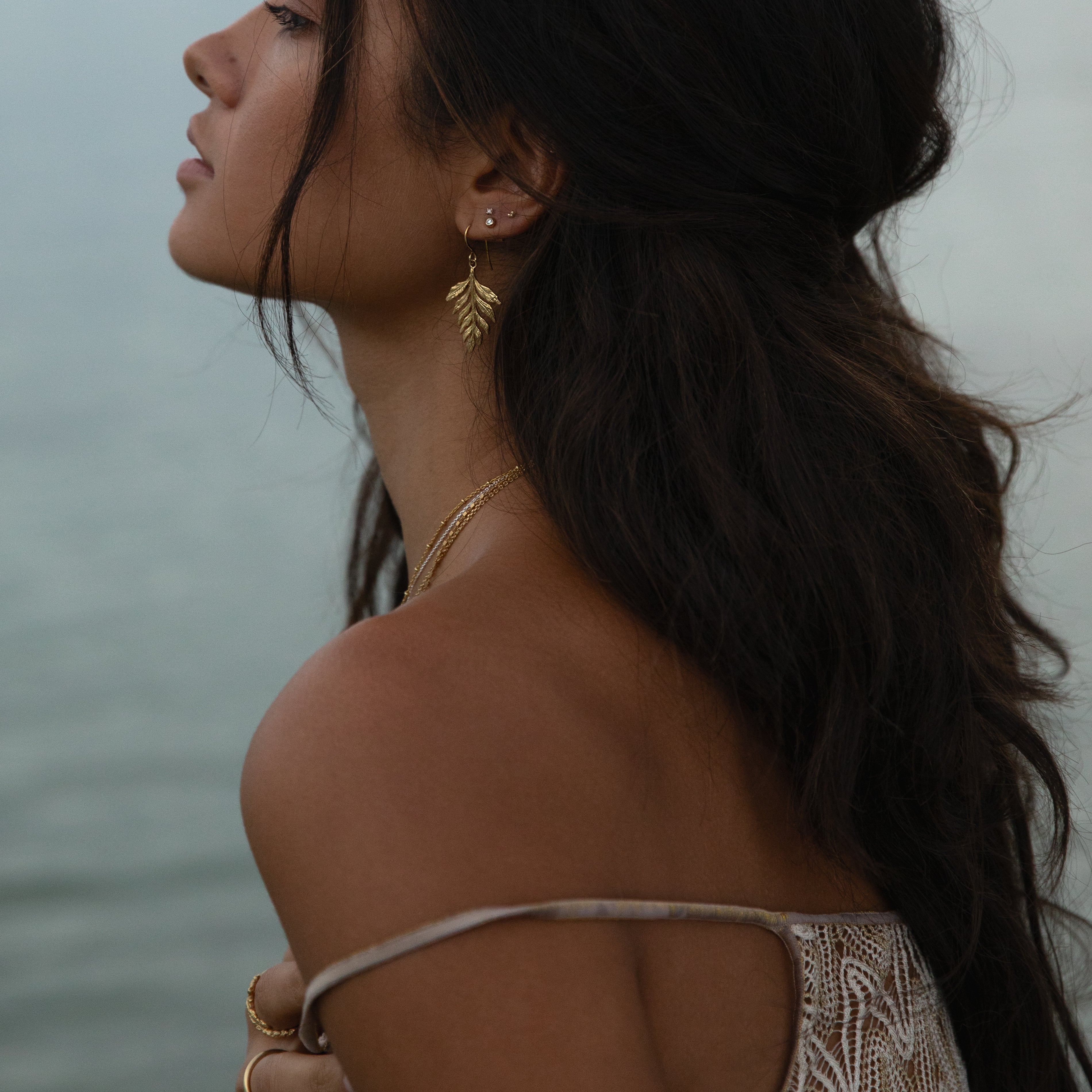 Gold fern drop earrings worn by model by the water, showcasing their elegant design and perfect for any occasion.