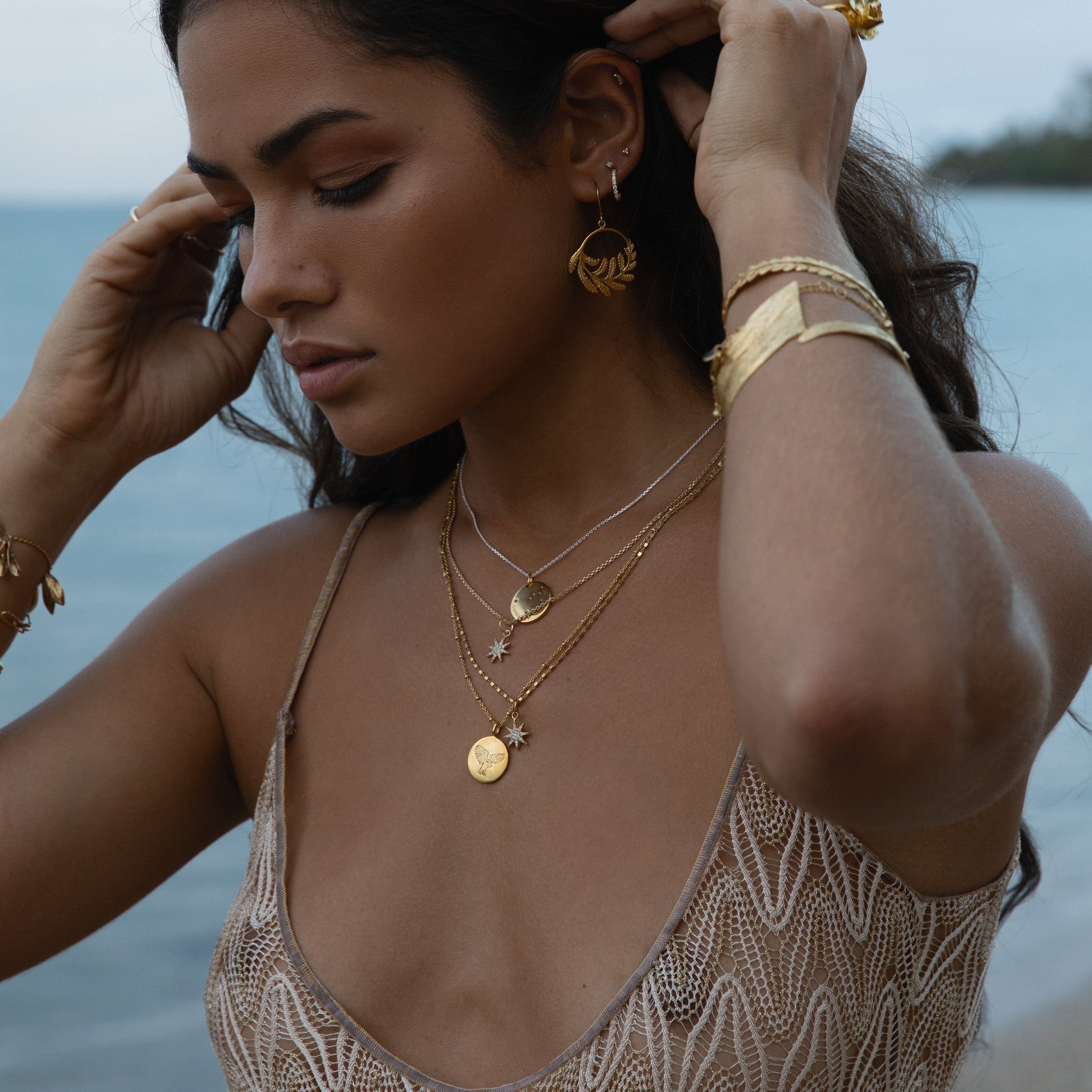 Model wearing gold fern hoop earrings with layered gold necklaces by the beach.
