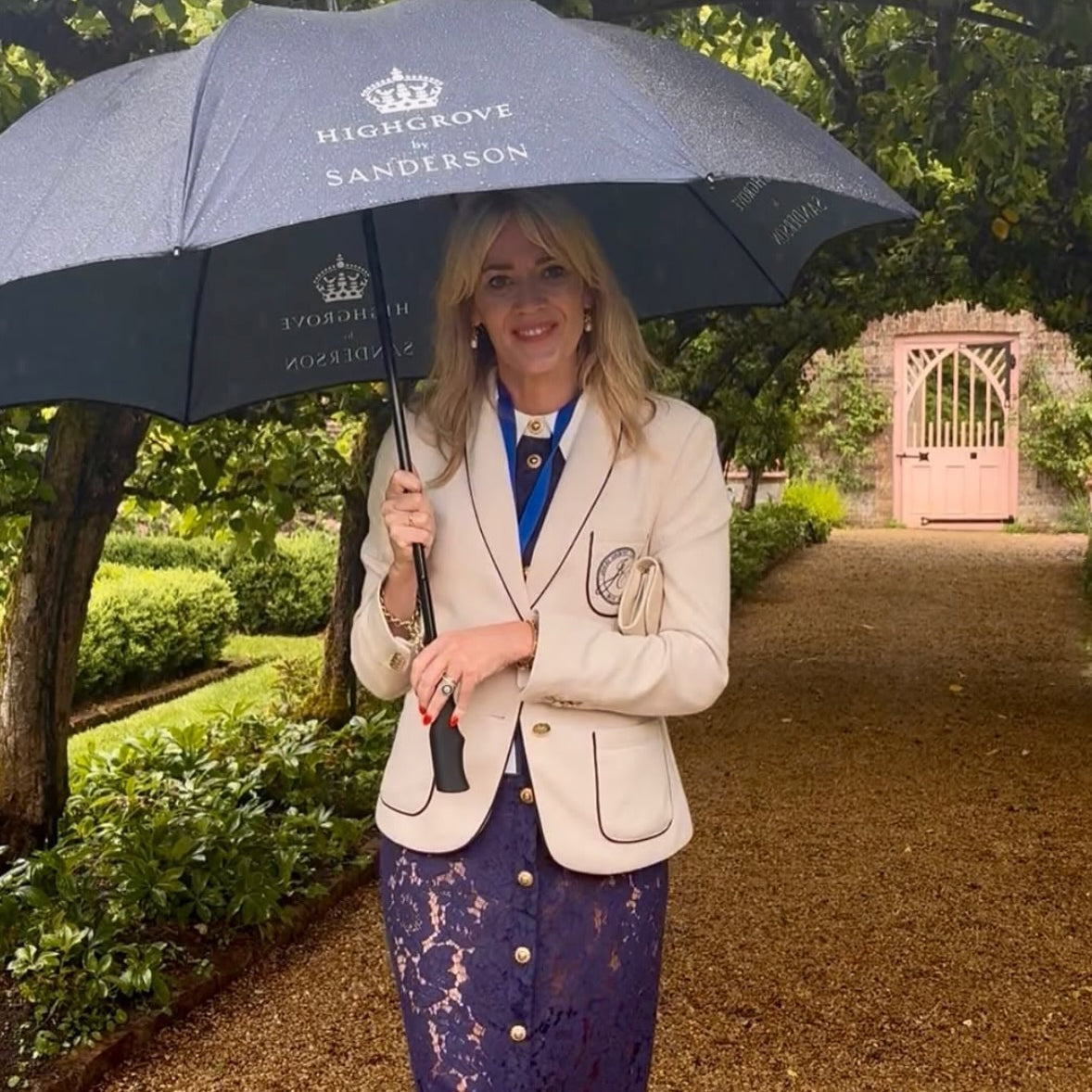 A woman stands on a garden path holding a dark navy umbrella with 'Highgrove by Sanderson' printed on it. She is wearing a cream blazer with piping, a blue top, and a purple lace skirt.