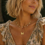 A woman with a short blonde bob is shown in a close-up portrait outdoors, wearing layered gold necklaces including a delicate chain dotted with cubic zirconia and a small butterfly pendant. She is wearing a floral-patterned top with ruffled shoulders.
