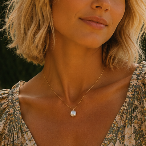 A woman wearing pearl necklace on a delicate gold chain, a sleeveless floral dress with ruffled shoulders and a deep V-neckline, standing outdoors in front of a stone building and garden plants.