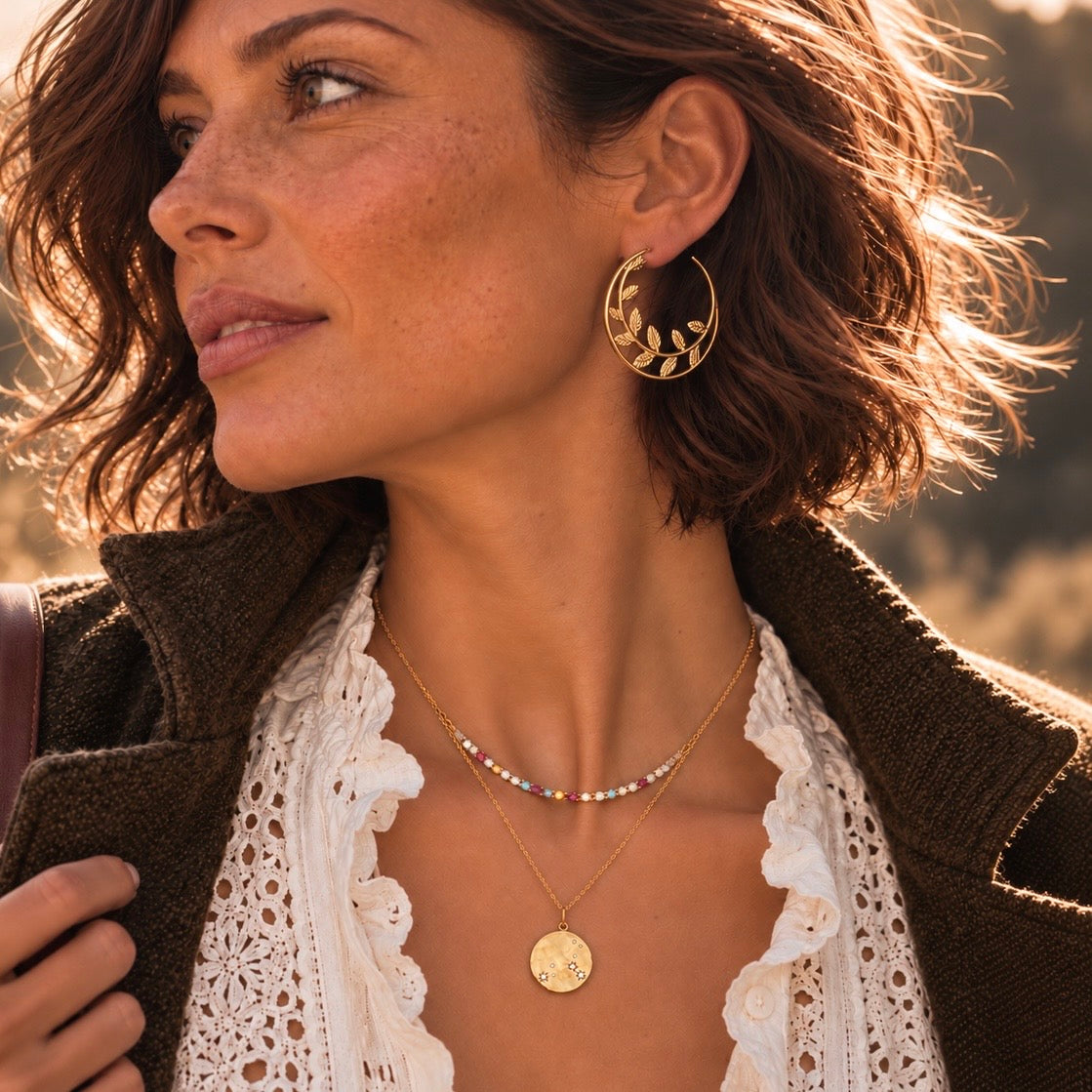 A woman in profile wearing gold hoop earrings with a leaf pattern, layered gold necklaces, and a white lace top under a brown jacket, in warm golden-hour sunlight.