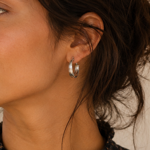 A woman with dark hair pulled back wears a mid sized silver hoop earring with a blade of grass texture, shown in a warm indoor setting. She is wearing a dark floral blouse.