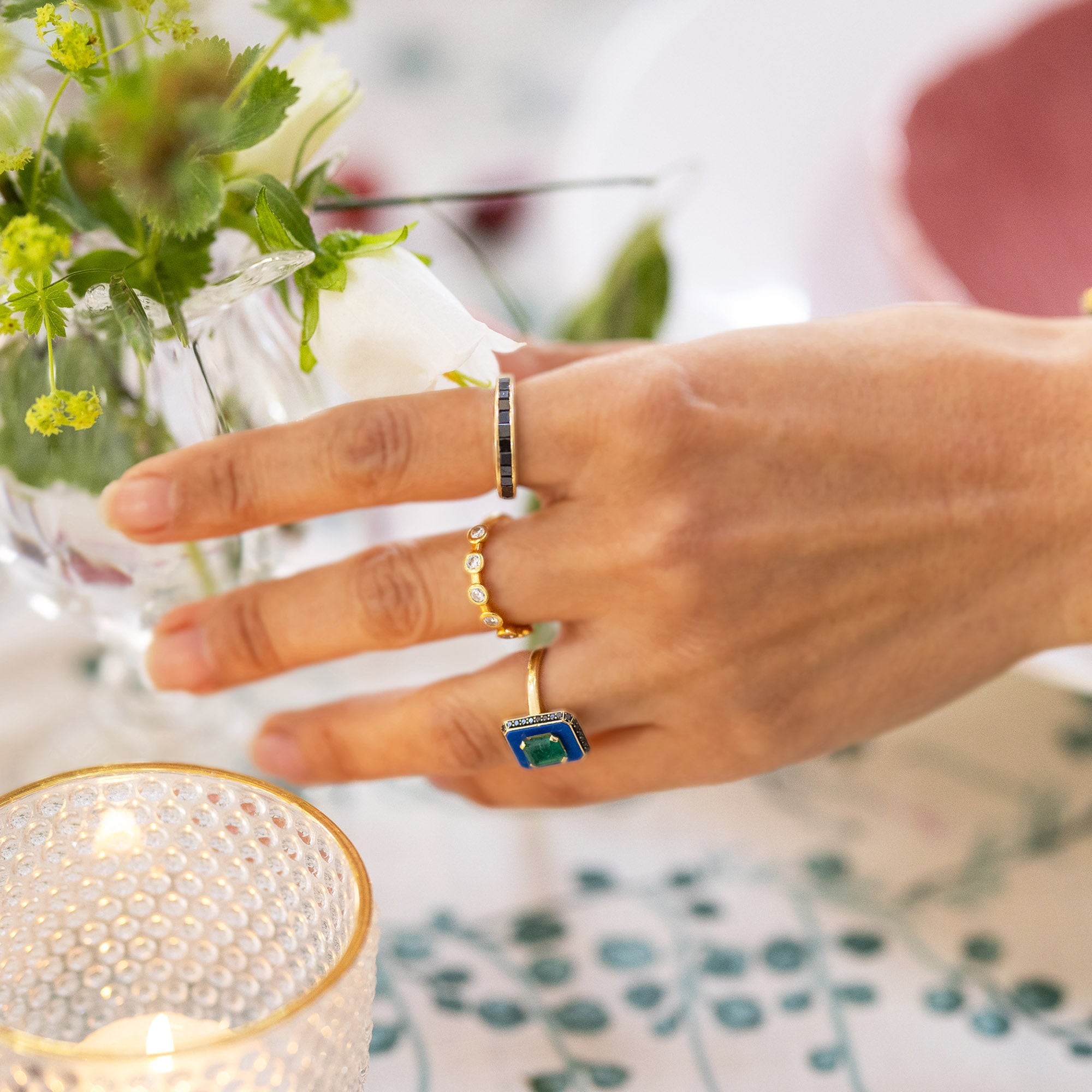 Close-up of a hand wearing the Gold Star Dust Ring among other rings, with a floral centerpiece and candle in the background.