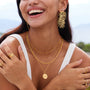Model wearing gold vetch earrings along with layered gold necklaces, smiling against a scenic backdrop.