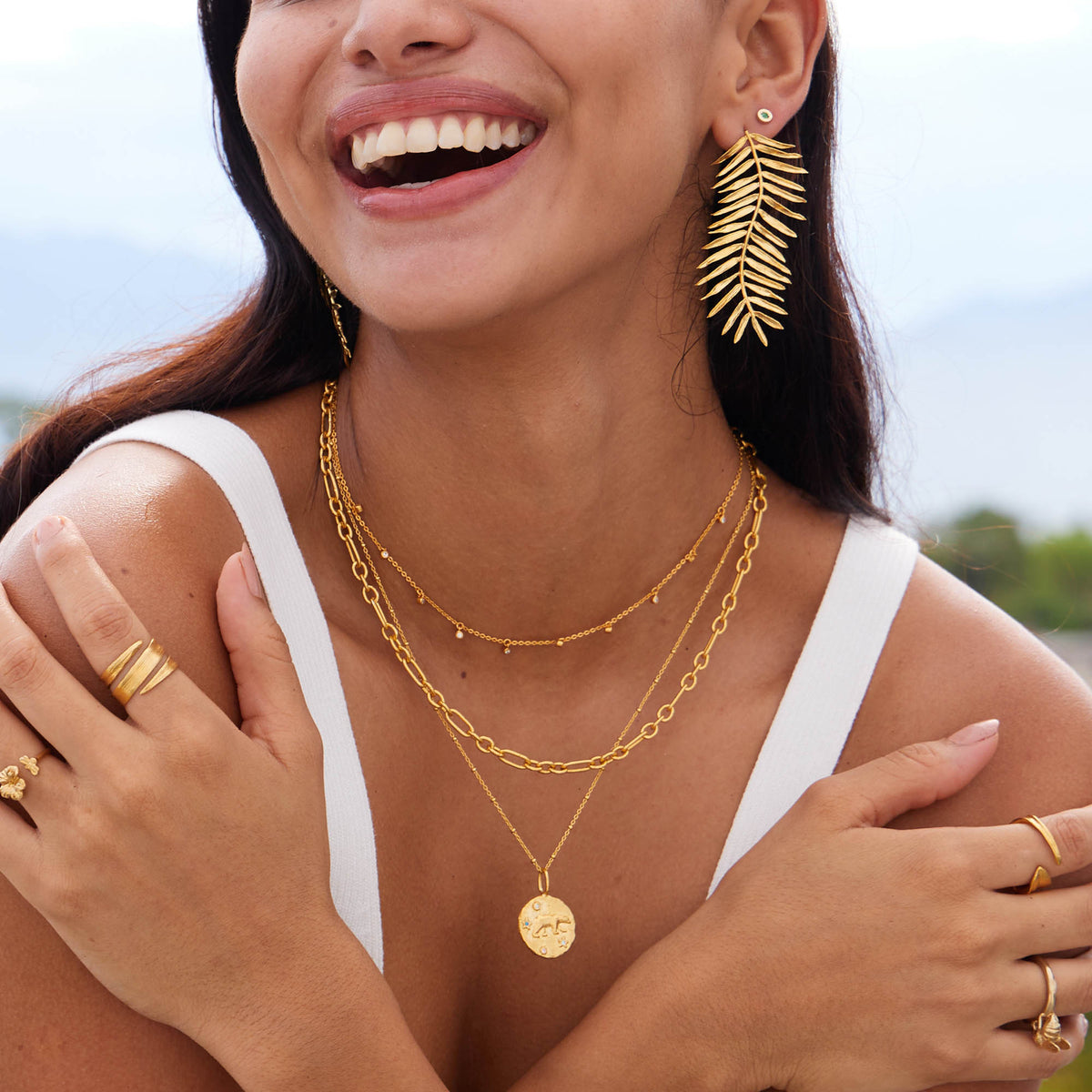 Model wearing gold vetch earrings along with layered gold necklaces, smiling against a scenic backdrop.