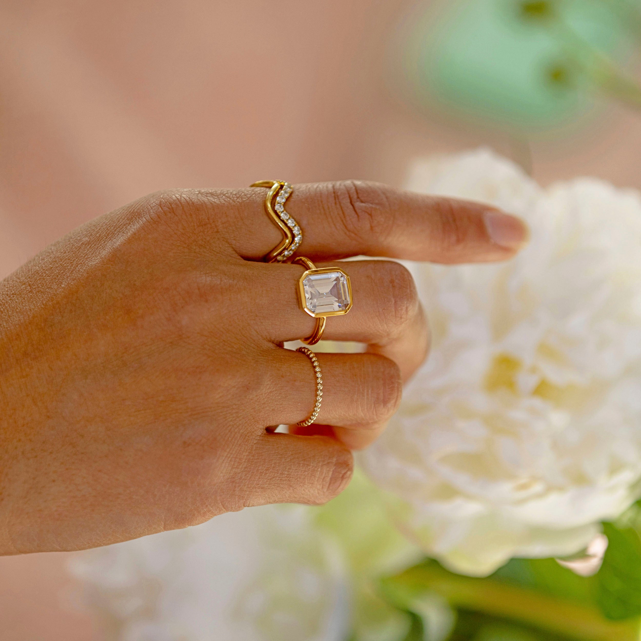 A hand adorned with a gold wave ring, showcasing elegant jewelry pieces alongside a blooming flower.