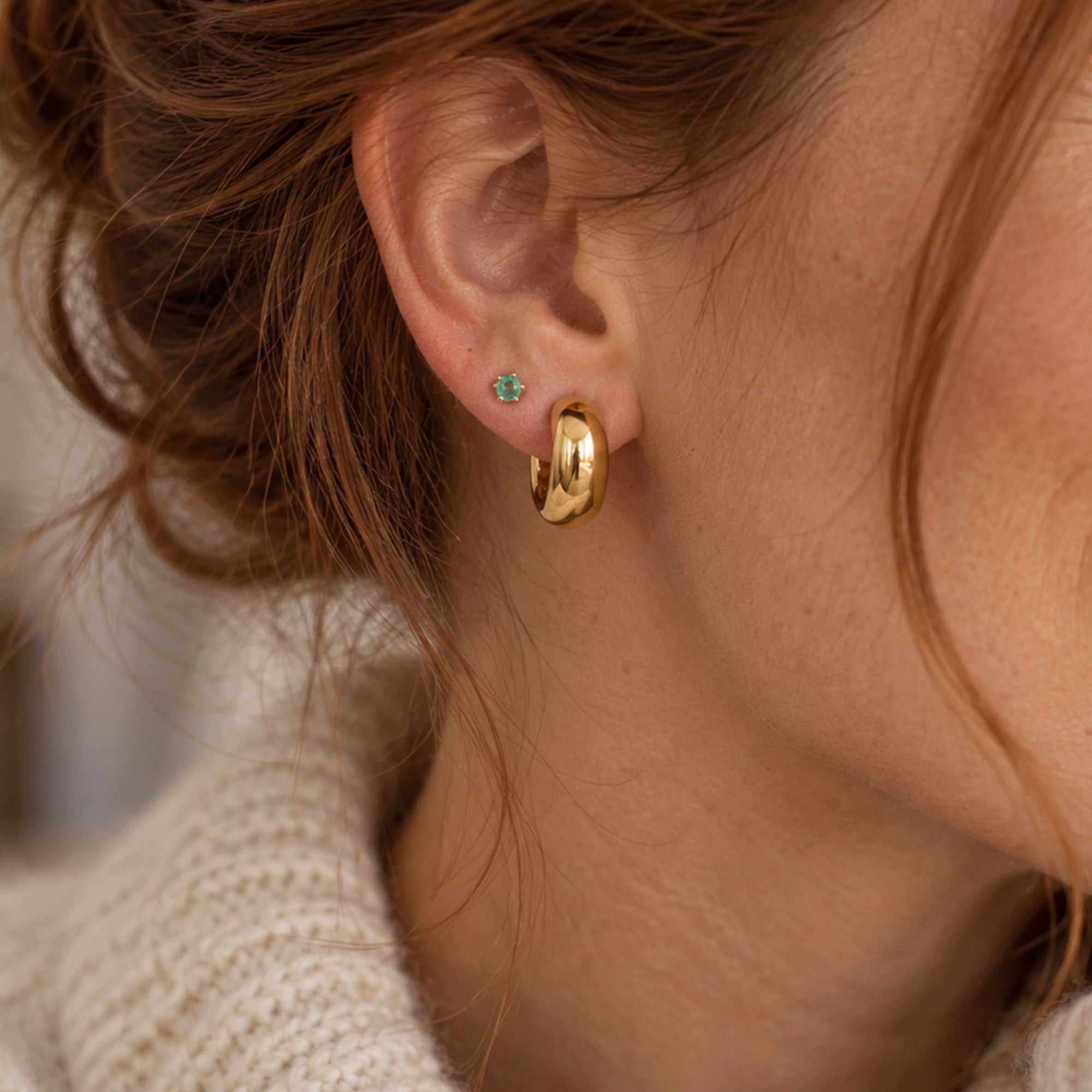 A close-up of a person's ear wearing a polished gold huggie hoop earring and a small green emerald gemstone stud earring, with auburn brown hair and a cream knit sweater visible.