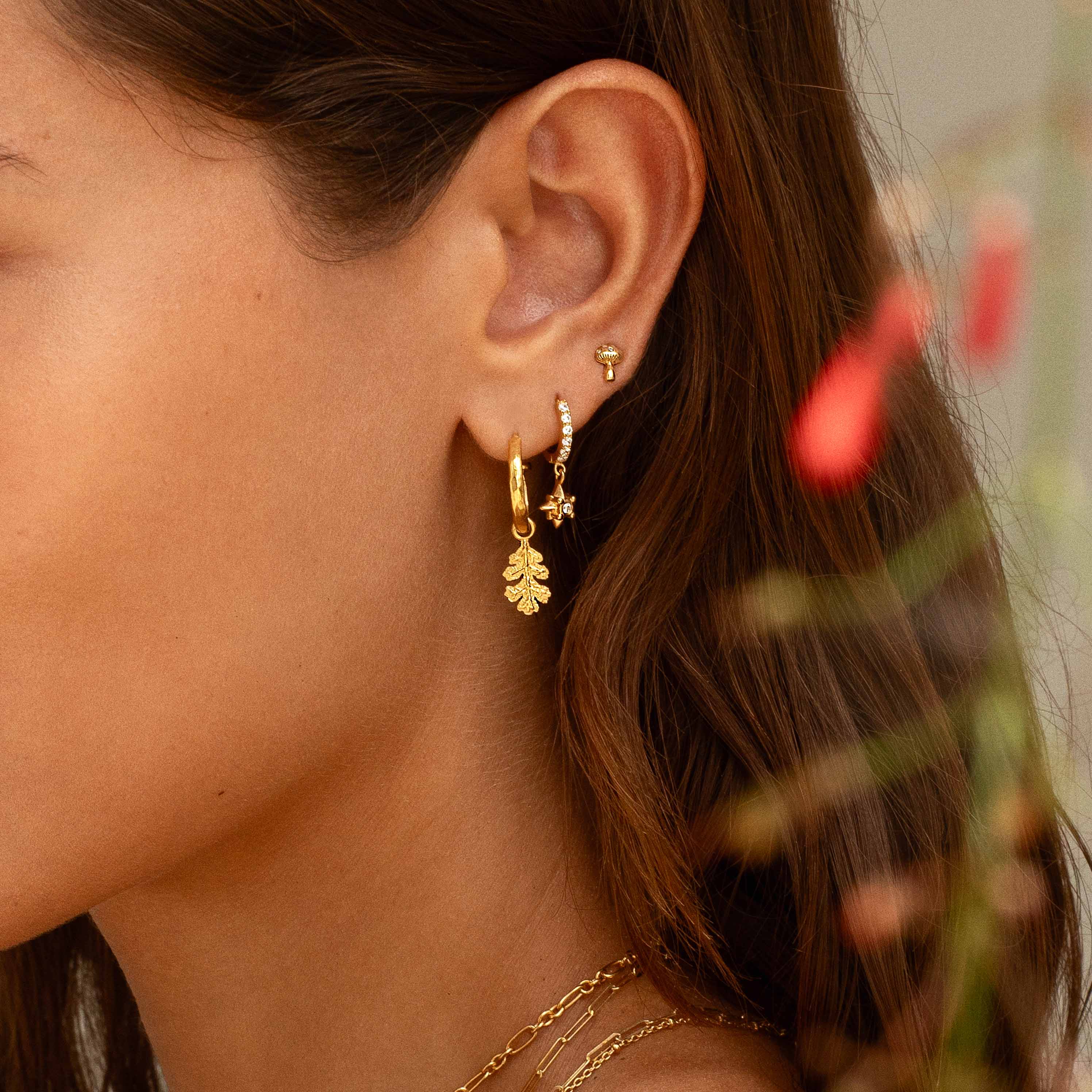 A close-up of a woman's ear wearing a gold huggie hoop earring with a dangling leaf charm and a small star charm, along with a tiny stud earring.