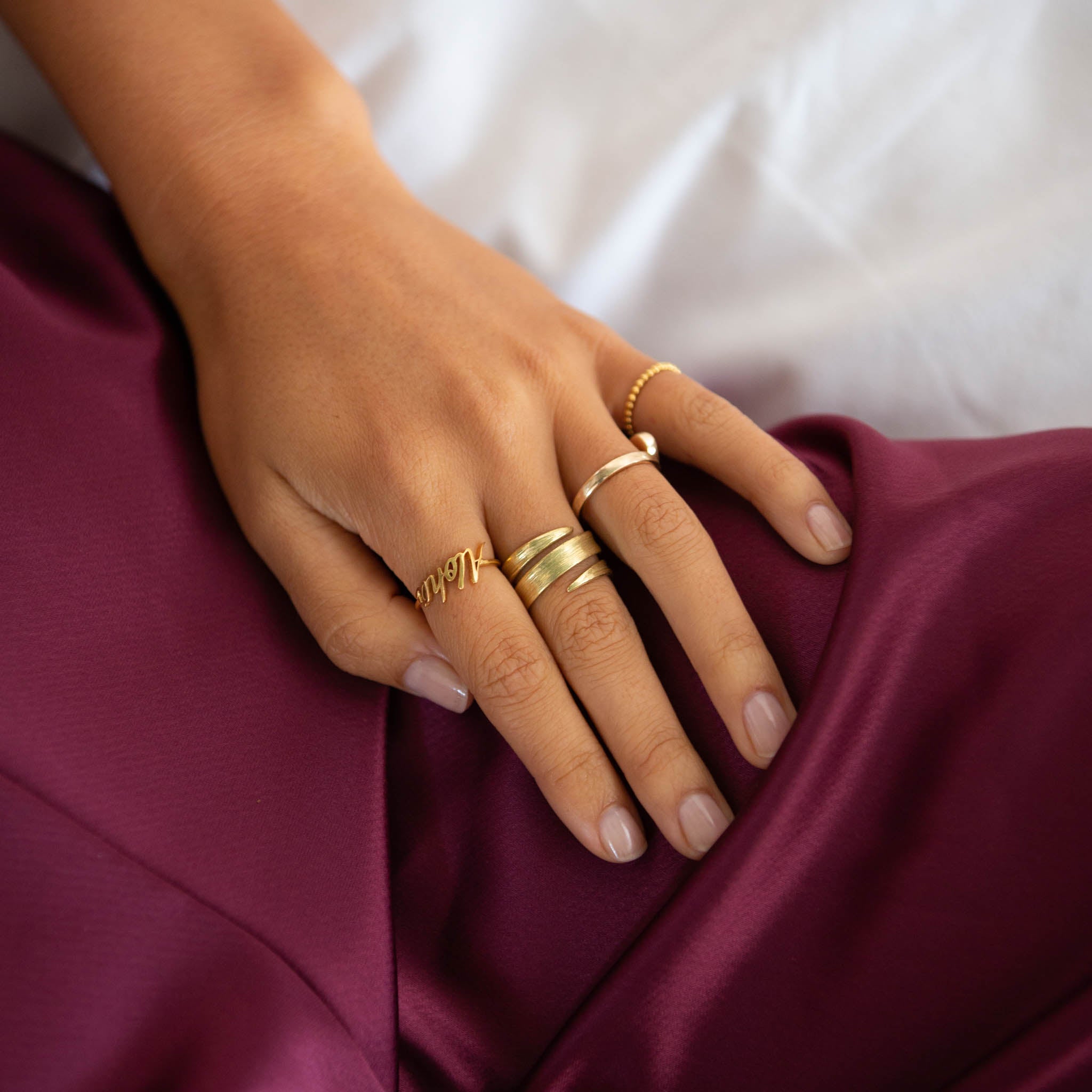 18ct solid gold blade of grass ring elegantly worn among other gold rings on a hand over a burgundy fabric.